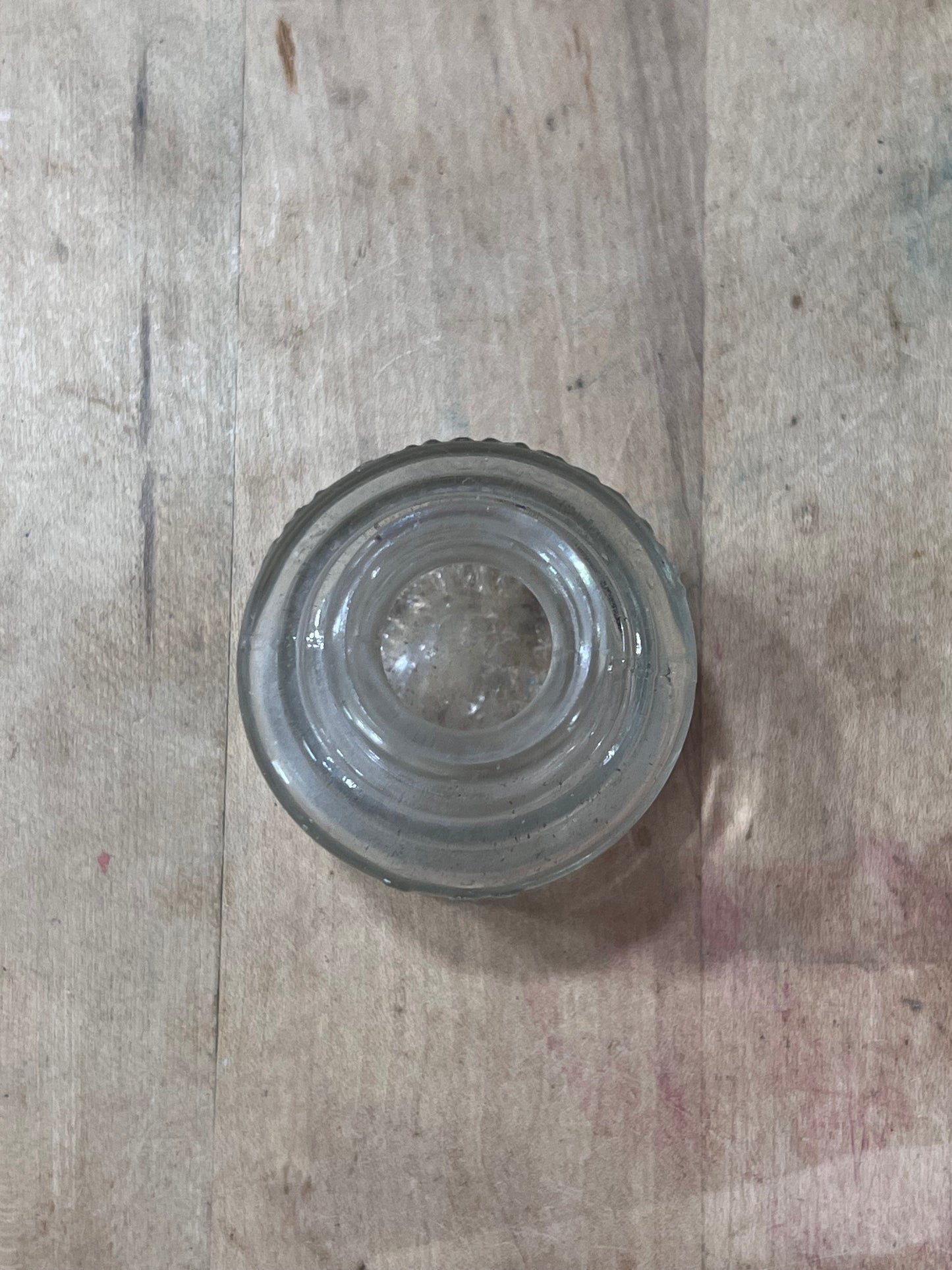 A clear glass bottle stopper with concentric circular ridges and a flared rim, shown from a top-down view on a light-colored wooden surface.