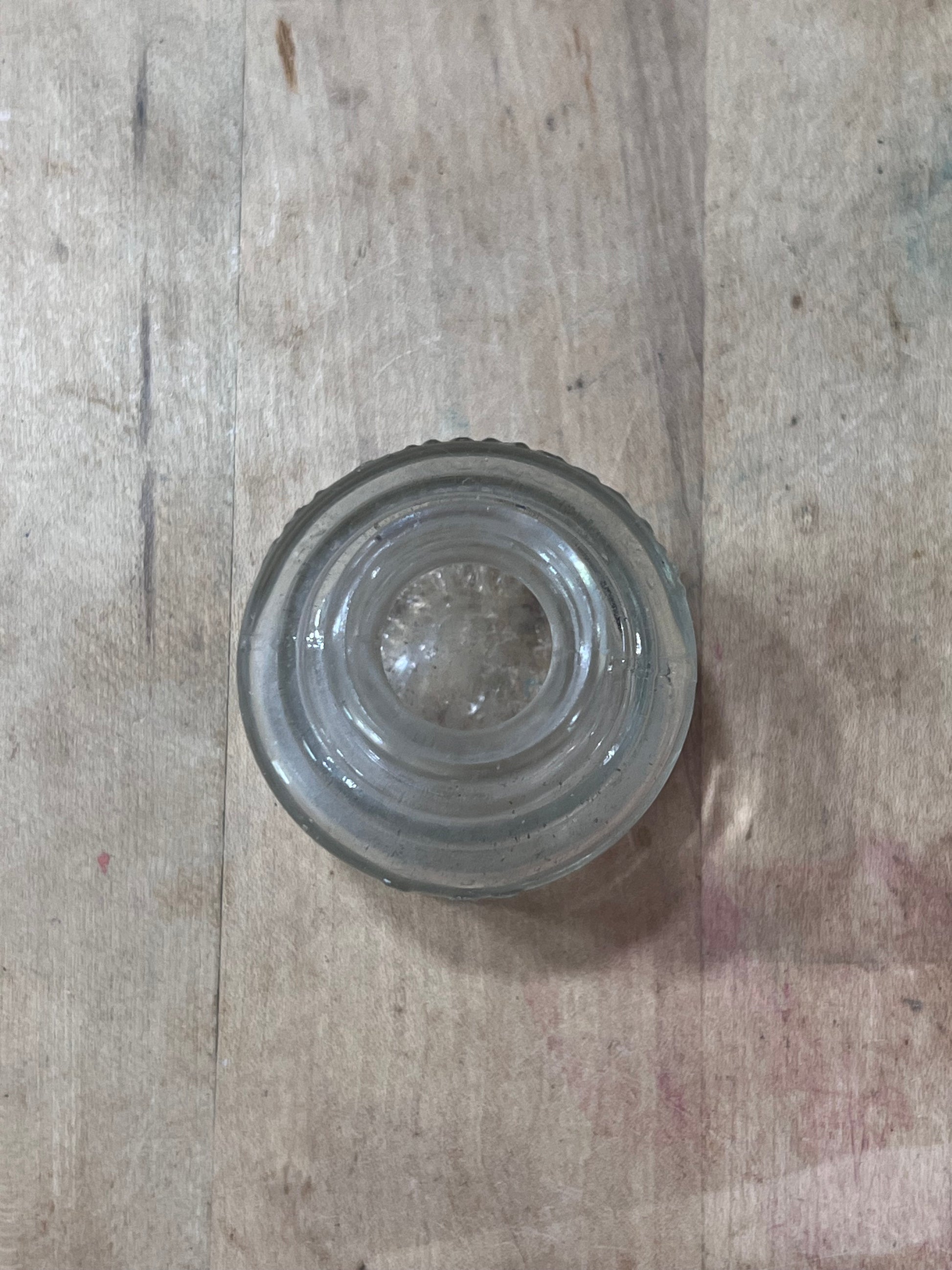 A clear glass bottle stopper with concentric circular ridges and a flared rim, shown from a top-down view on a light-colored wooden surface.