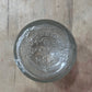 A clear glass jar lid with embossed vintage branding, photographed from directly above on a light wood surface.