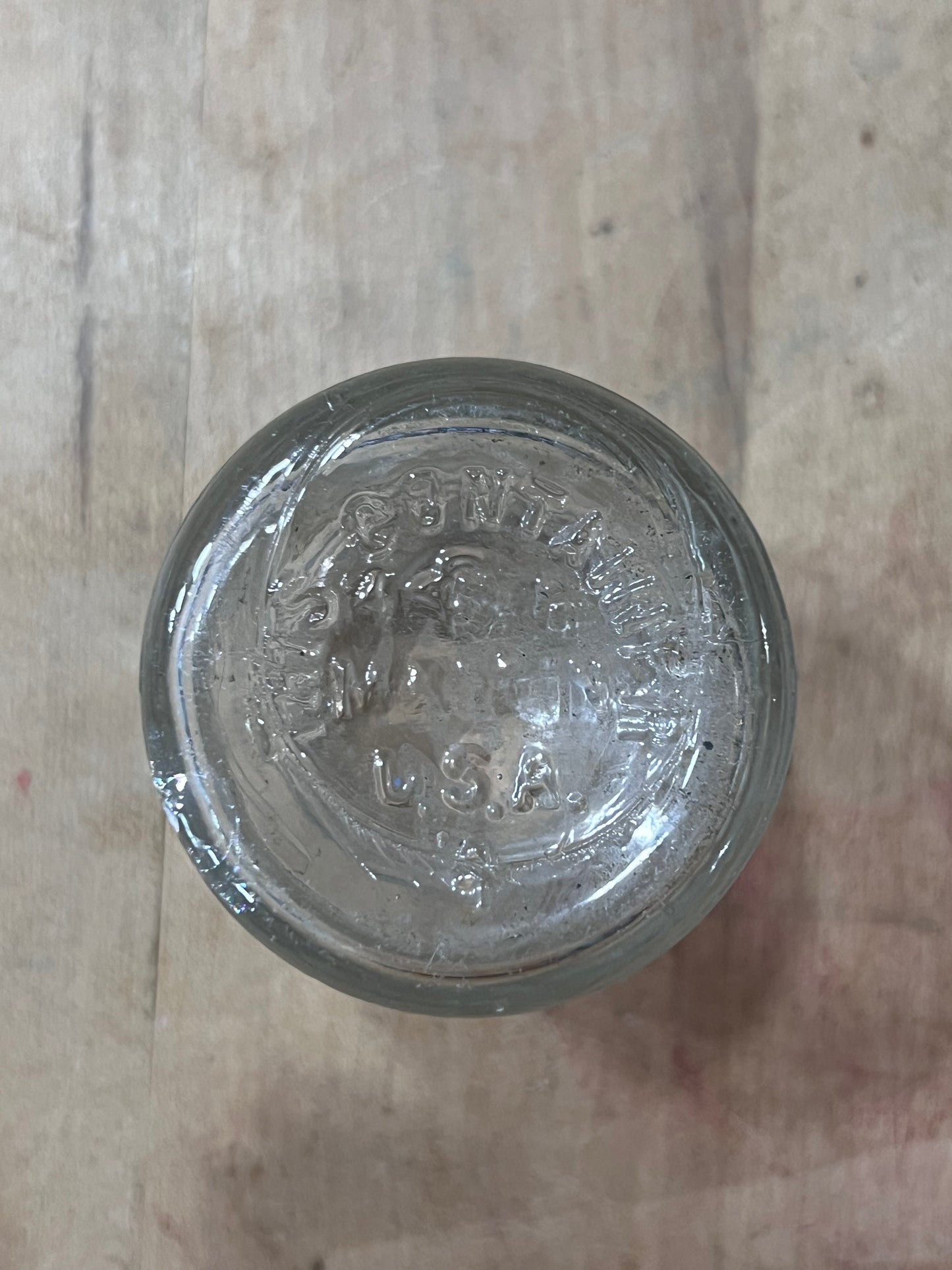 A clear glass jar lid with embossed vintage branding, photographed from directly above on a light wood surface.