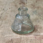 A clear glass bottle stopper or small glass container with a flared base, photographed on a worn beige-brown wooden surface.