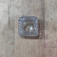 A small, clear textured glass ashtray with a square shape and wavy edges, resting on a light brown wooden surface.