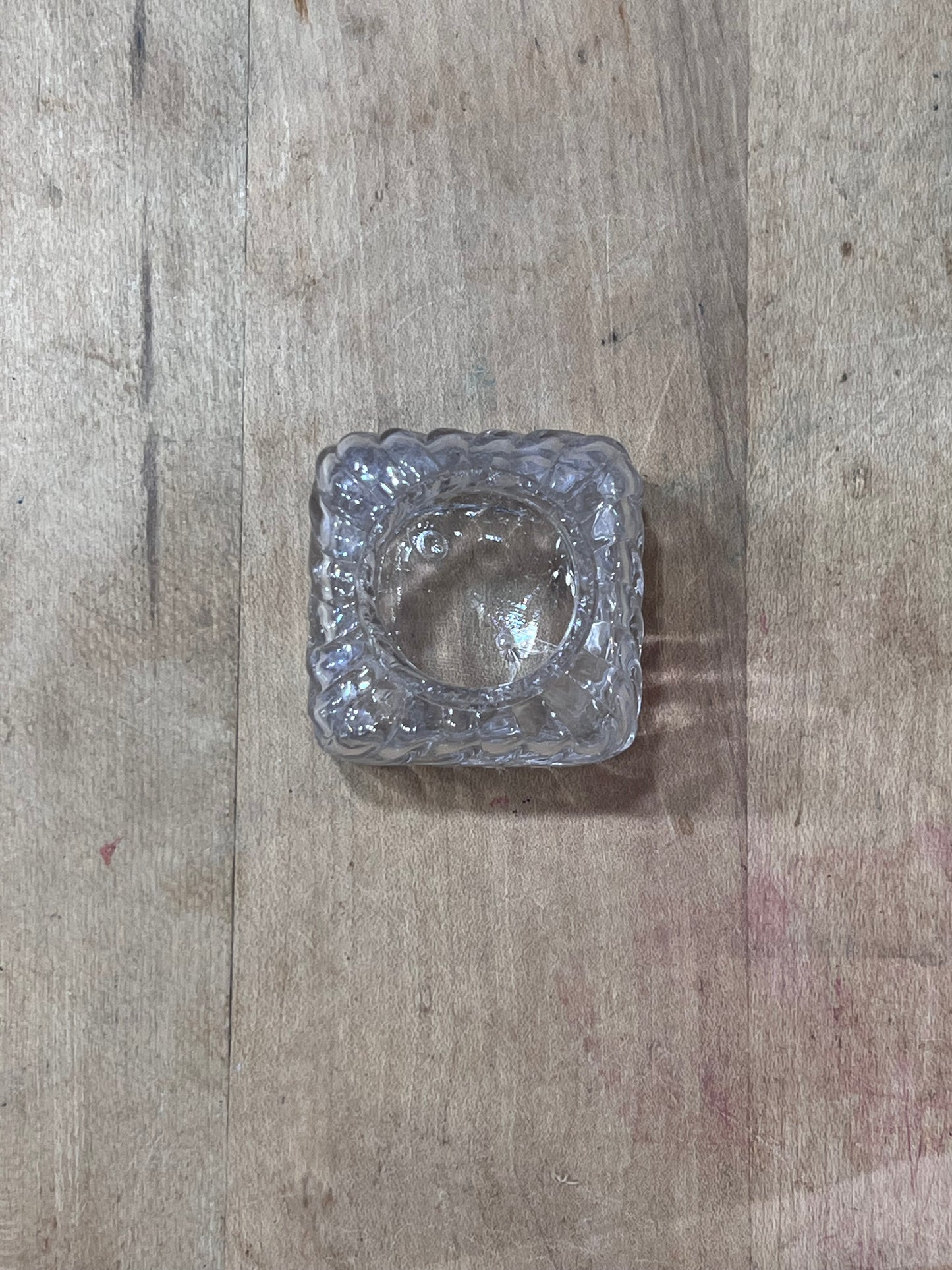 A small, clear textured glass ashtray with a square shape and wavy edges, resting on a light brown wooden surface.