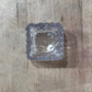 A small square clear glass ashtray with a scalloped edge, photographed from above on a light brown wooden surface.