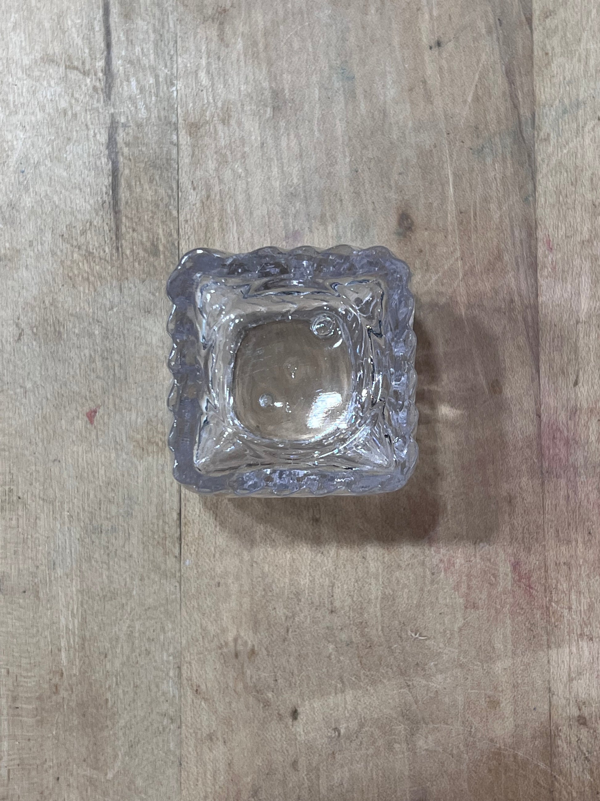 A small square clear glass ashtray with a scalloped edge, photographed from above on a light brown wooden surface.
