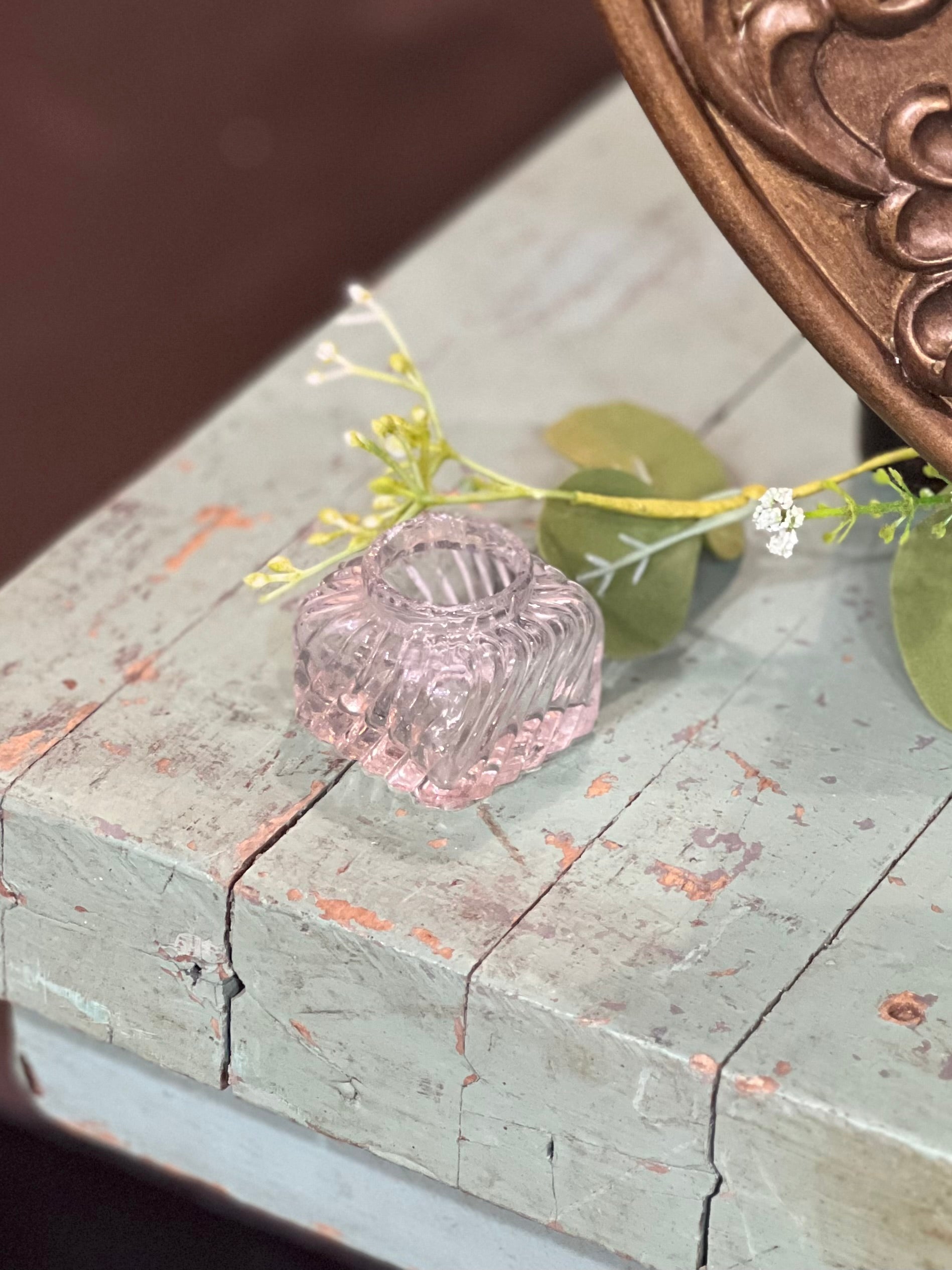 A small, clear glass candle holder with a textured, ribbed pattern sits on a distressed, light blue wooden surface decorated with green leaves and white flowers.