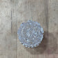 A round, faceted clear glass or crystal dish with a textured, cut pattern, resting on a light-colored wooden surface.