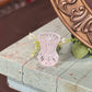 A small, pale pink cut-crystal vase with a textured, openwork pattern, displayed on a distressed light blue wooden table next to a large bronze mirror.