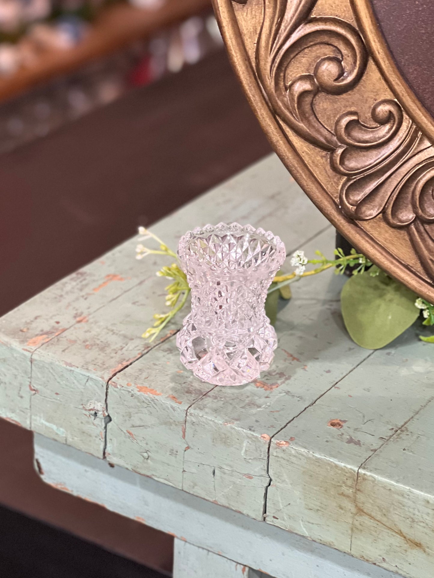 A small, pale pink cut-crystal vase with a textured, openwork pattern, displayed on a distressed light blue wooden table next to a large bronze mirror.