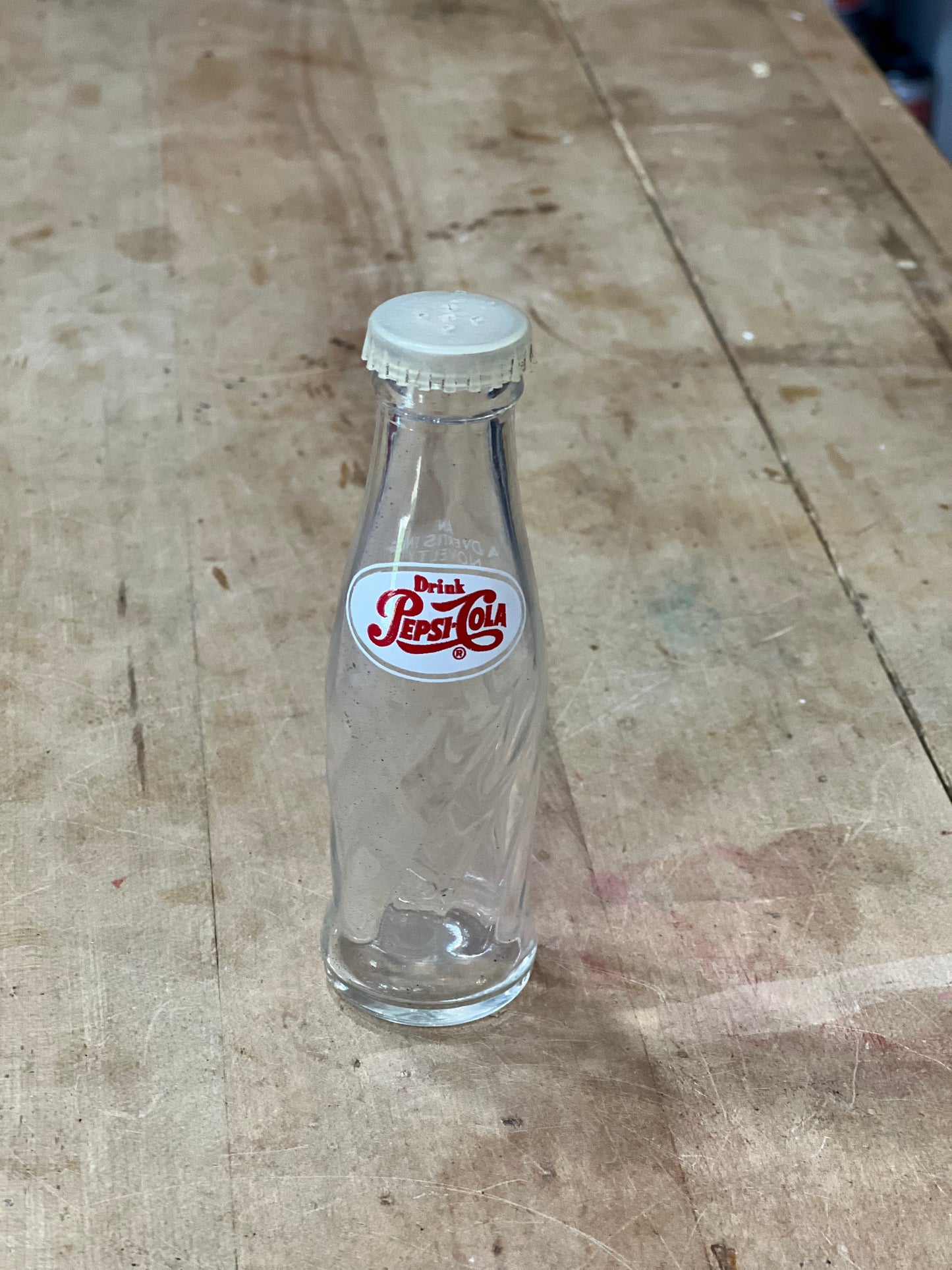 A clear glass bottle with a white screw cap and a white label featuring red Arabic script, lying on a worn wooden surface.