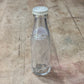 A clear glass bottle with a white cap, lying on a rustic wooden surface. The bottle features white printed text that reads 'A NEW MEAN GIRL MOVIE' alongside a circular logo.