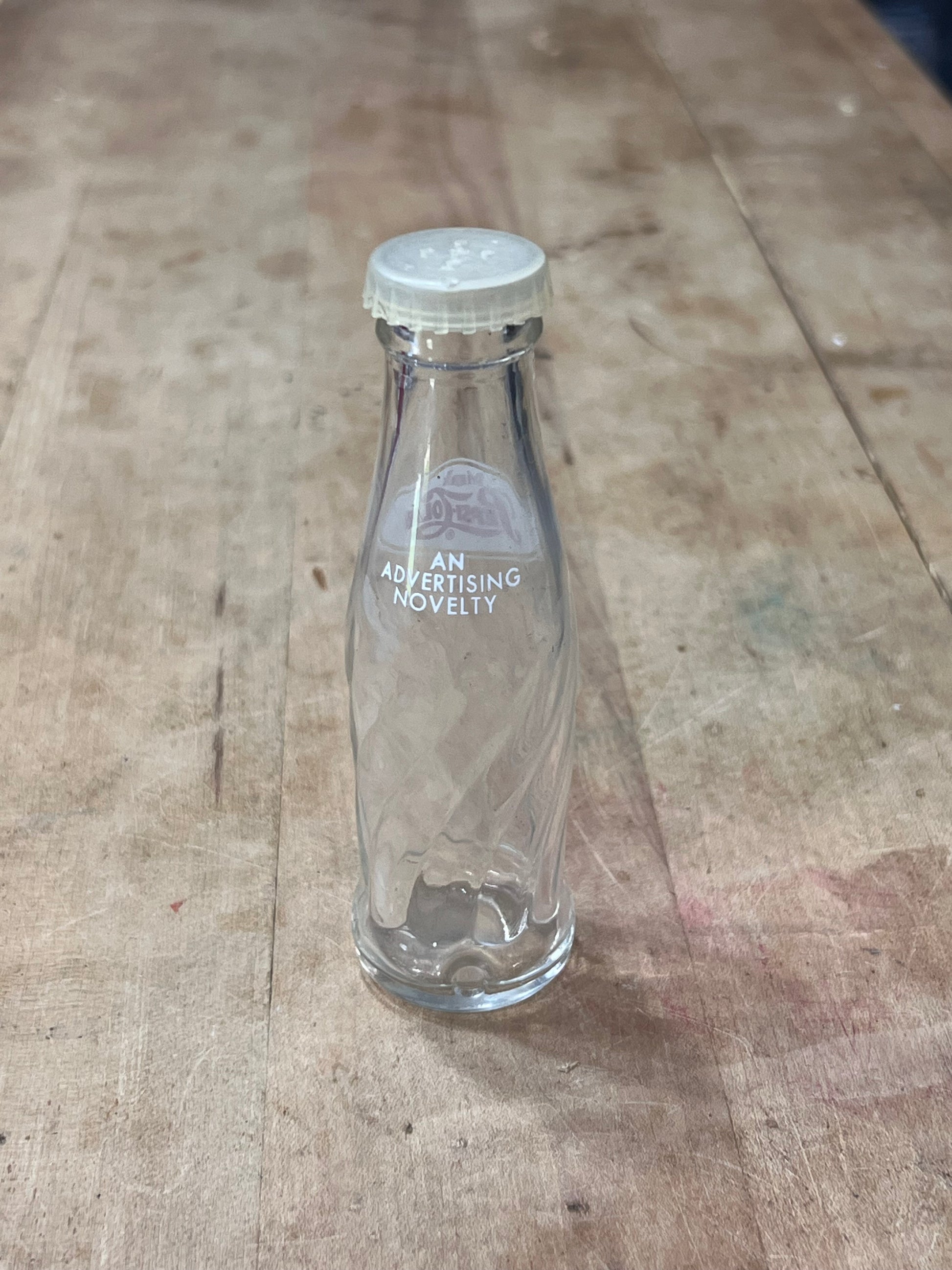 A clear glass bottle with a white cap, lying on a rustic wooden surface. The bottle features white printed text that reads 'A NEW MEAN GIRL MOVIE' alongside a circular logo.