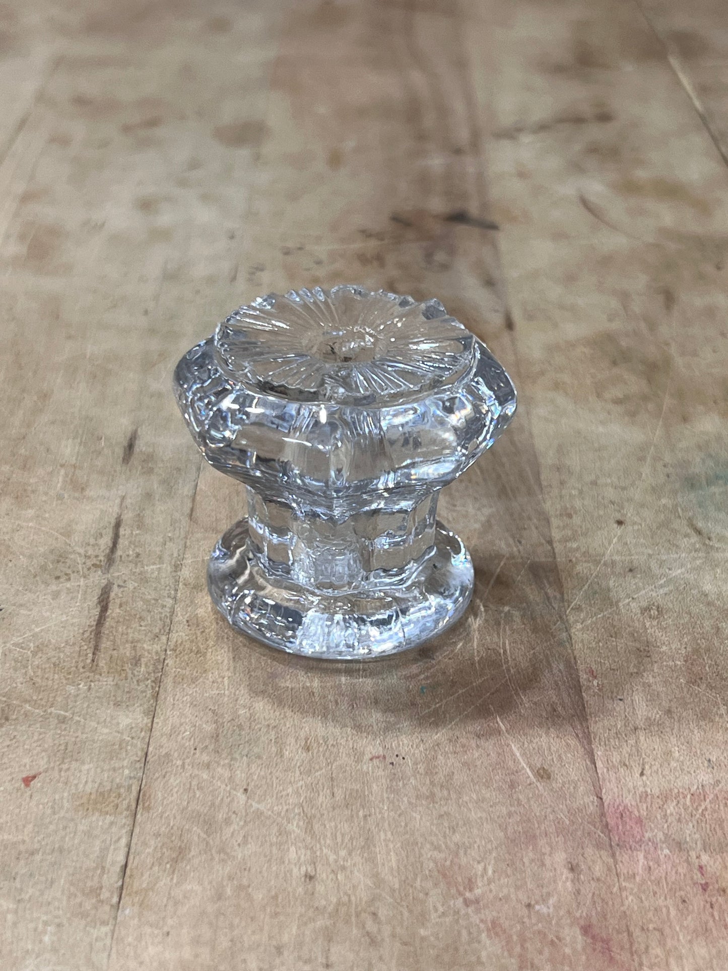 A clear glass cabinet knob with a faceted, ribbed pattern, resting on a light brown wooden surface.