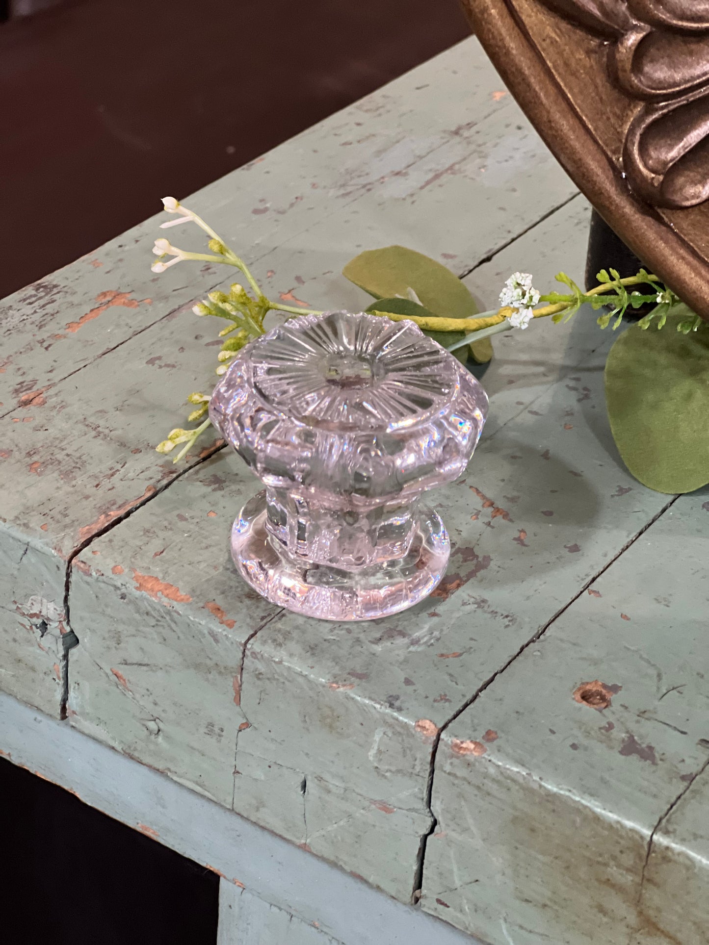 A vintage-style clear glass doorknob with a faceted, starburst pattern, mounted on a distressed, light blue-gray wooden cabinet with decorative greenery.