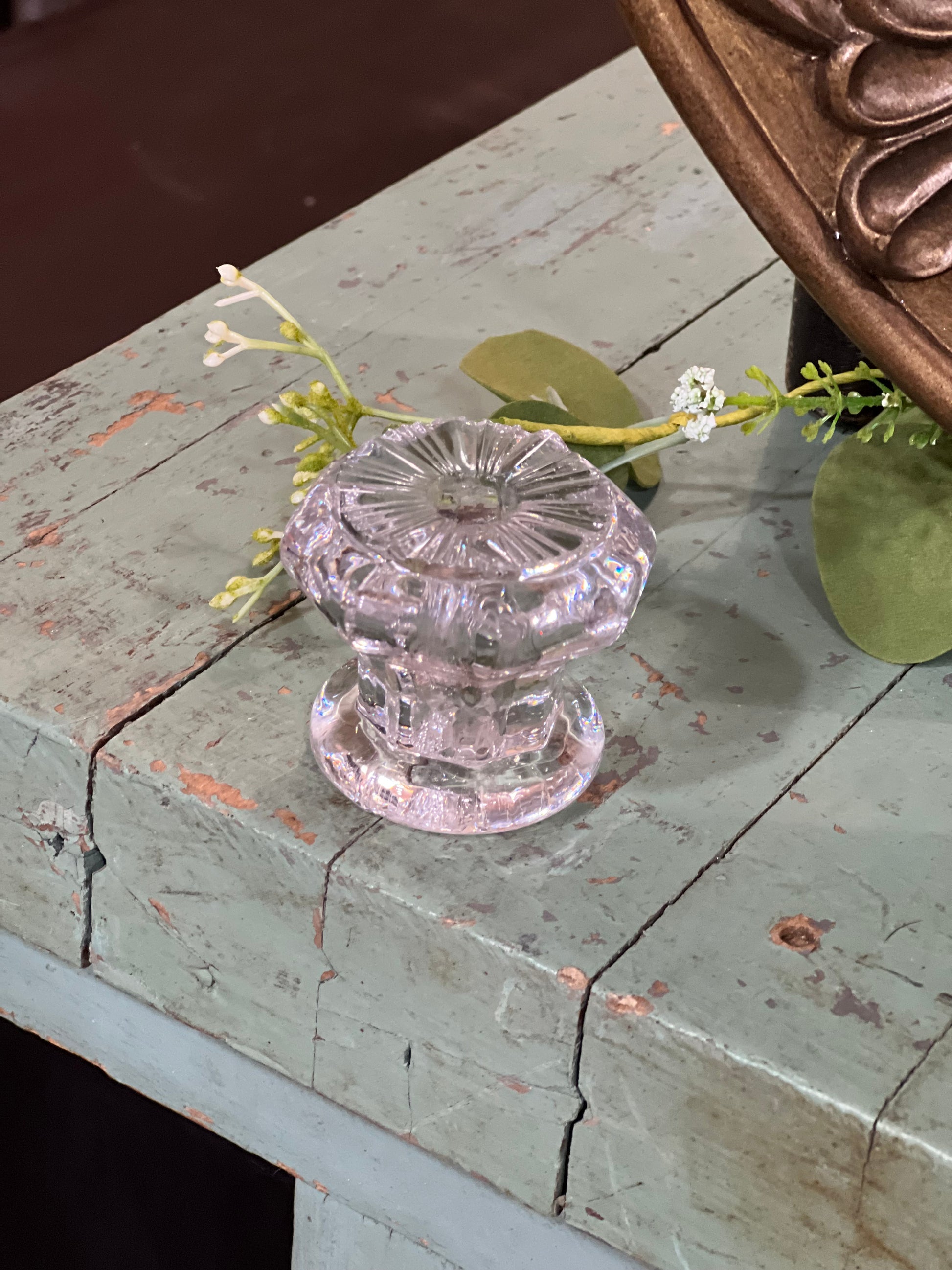 A vintage-style clear glass doorknob with a faceted, starburst pattern, mounted on a distressed, light blue-gray wooden cabinet with decorative greenery.