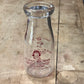 A vintage clear glass milk bottle with red printed text and a cartoon illustration, lying on a weathered wooden surface.