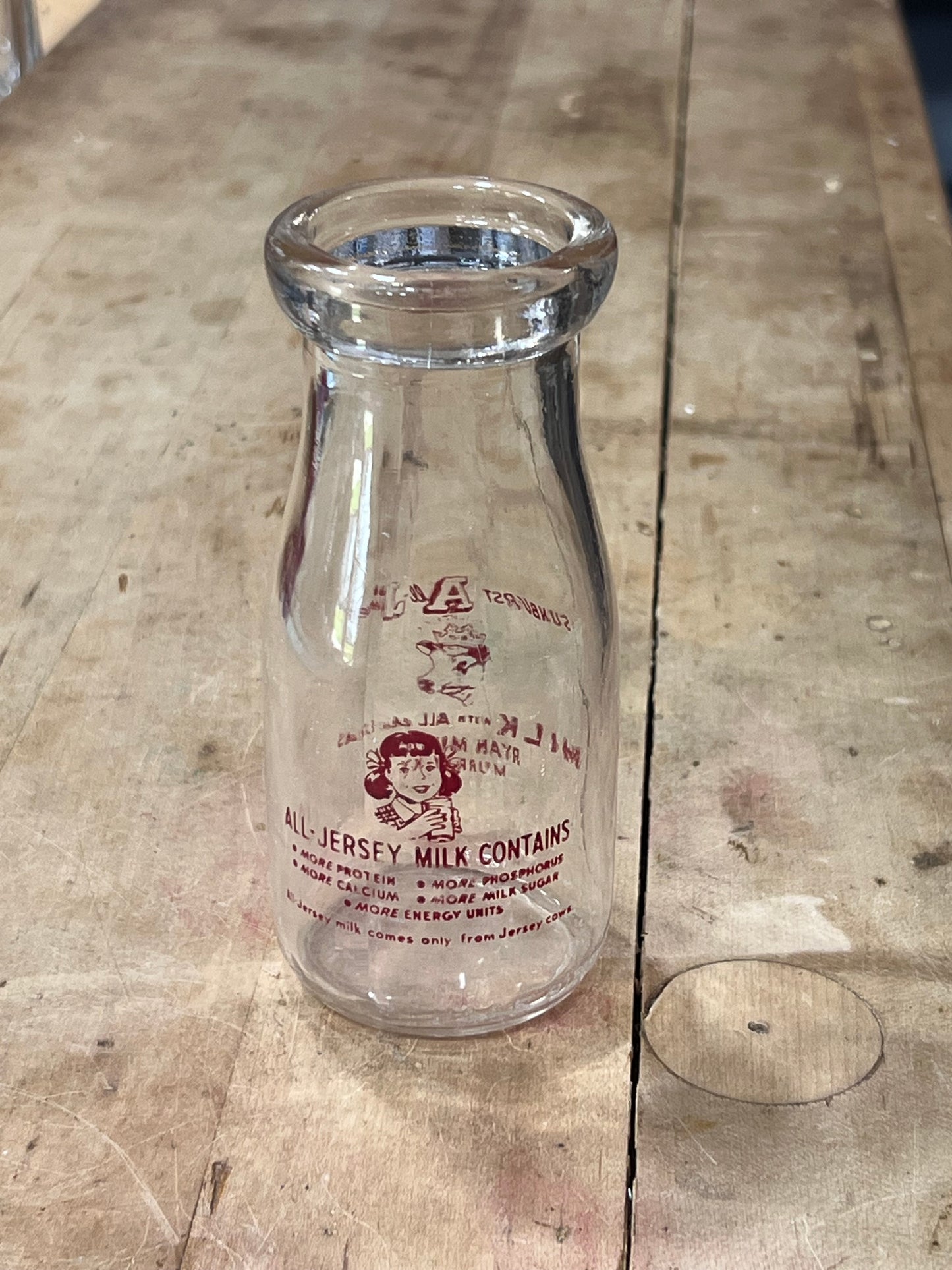 A vintage clear glass milk bottle with red printed text and a cartoon illustration, lying on a weathered wooden surface.