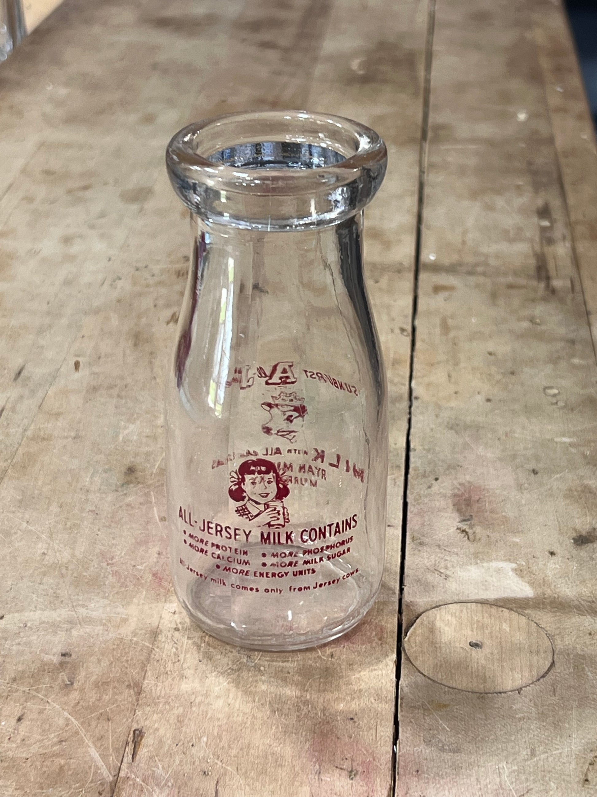 A vintage clear glass milk bottle with red printed text and a cartoon illustration, lying on a weathered wooden surface.