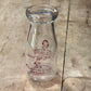 A clear glass milk bottle with red vintage printed branding, lying on a rustic wooden plank surface.