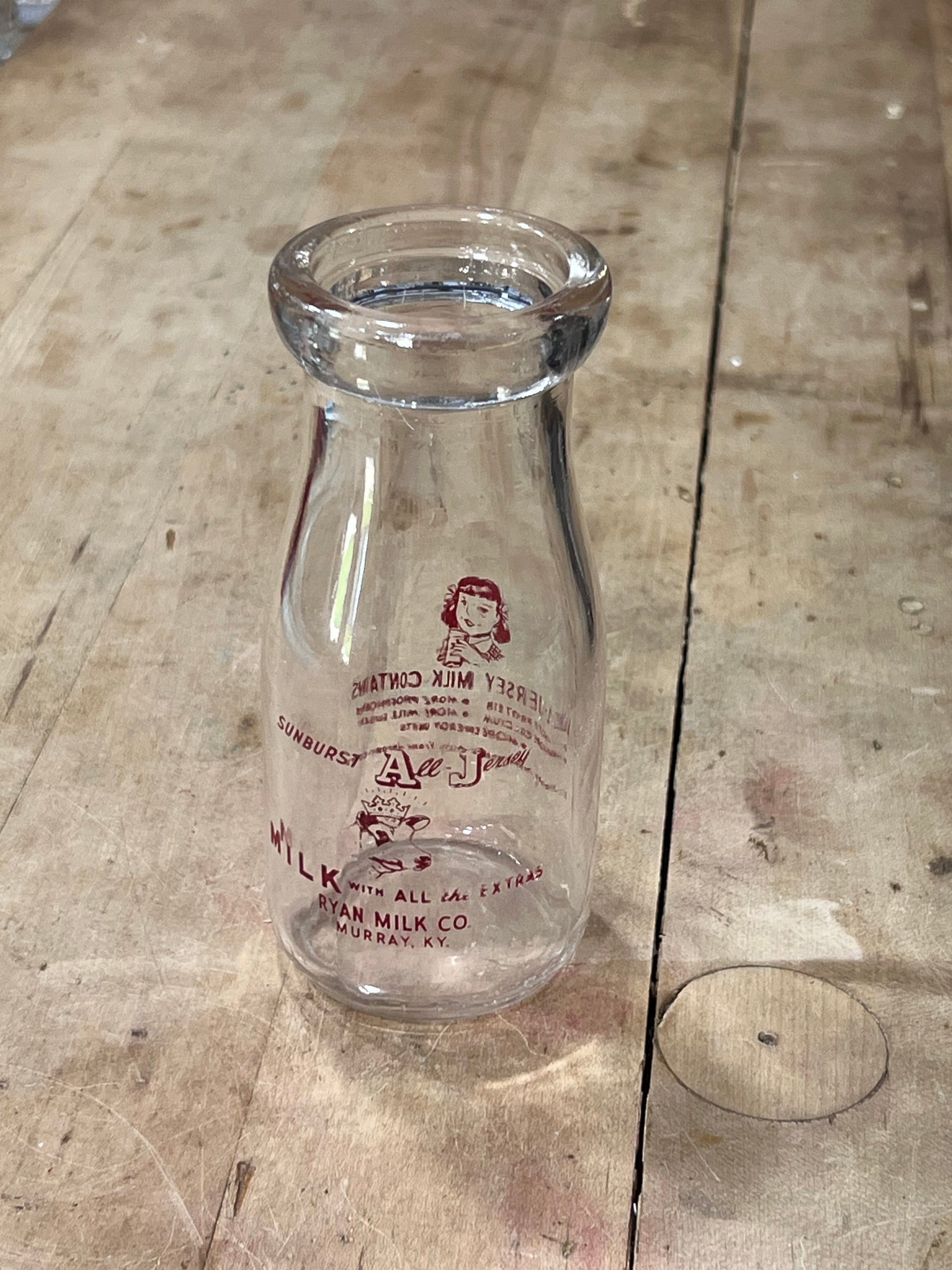 A clear glass milk bottle with red vintage printed branding, lying on a rustic wooden plank surface.