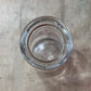 A clear glass bottle viewed from above, showing the circular glass opening and a small amount of white residue or sediment inside, set against a light gray textured background.
