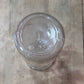 A clear glass jar or bottle viewed from above, showing the bottom of the container with embossed markings, placed on a light wood surface.