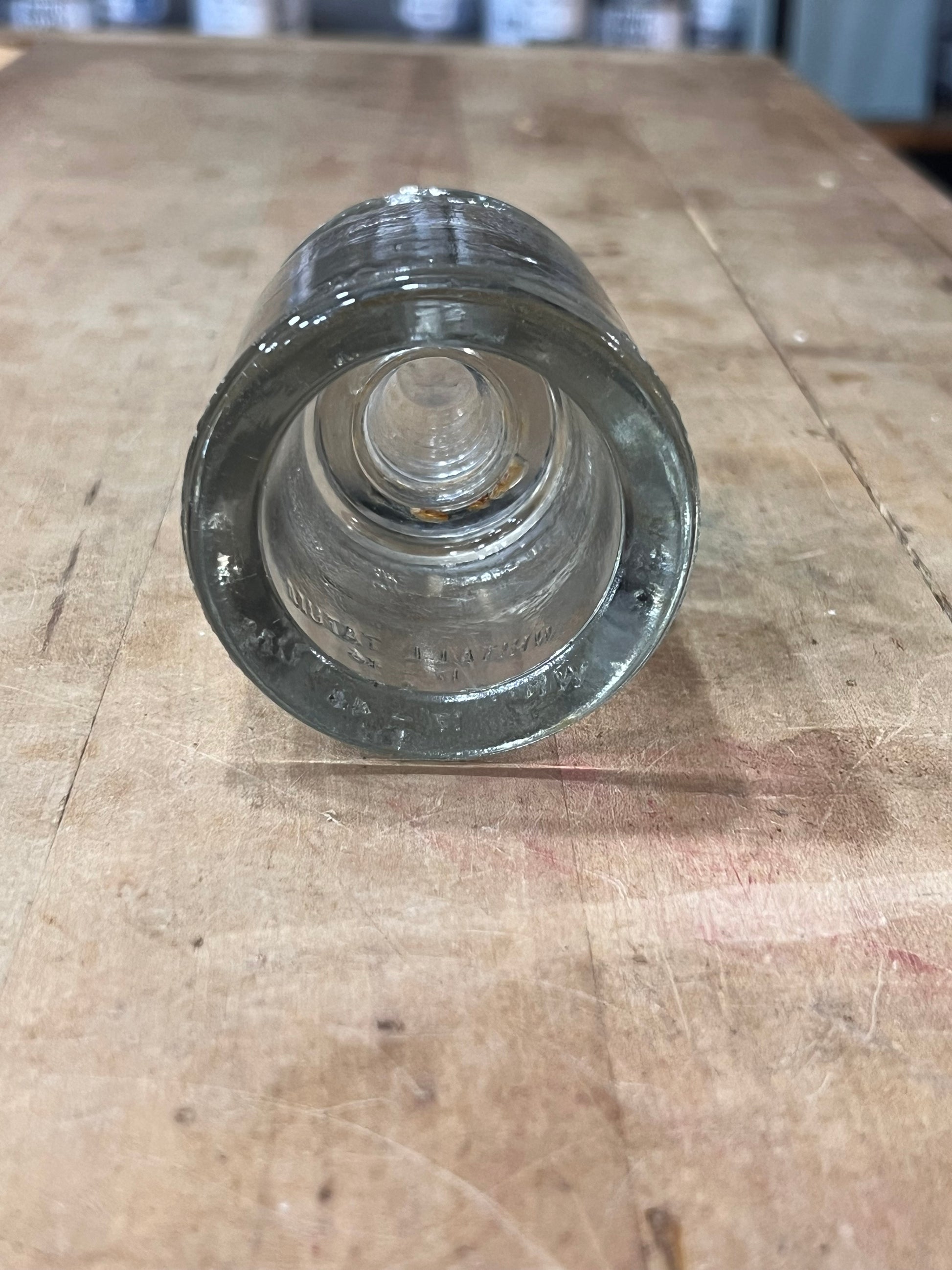 A clear glass bottle stopper or cap with a textured, ribbed outer ring and a smooth, domed center, shown from a top-down perspective on a worn wooden tabletop.