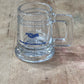 A clear glass beer mug with a handle, featuring a blue printed 'NCL' logo and 'Hawaiian Caribbean Lines' text, resting on a wooden surface.