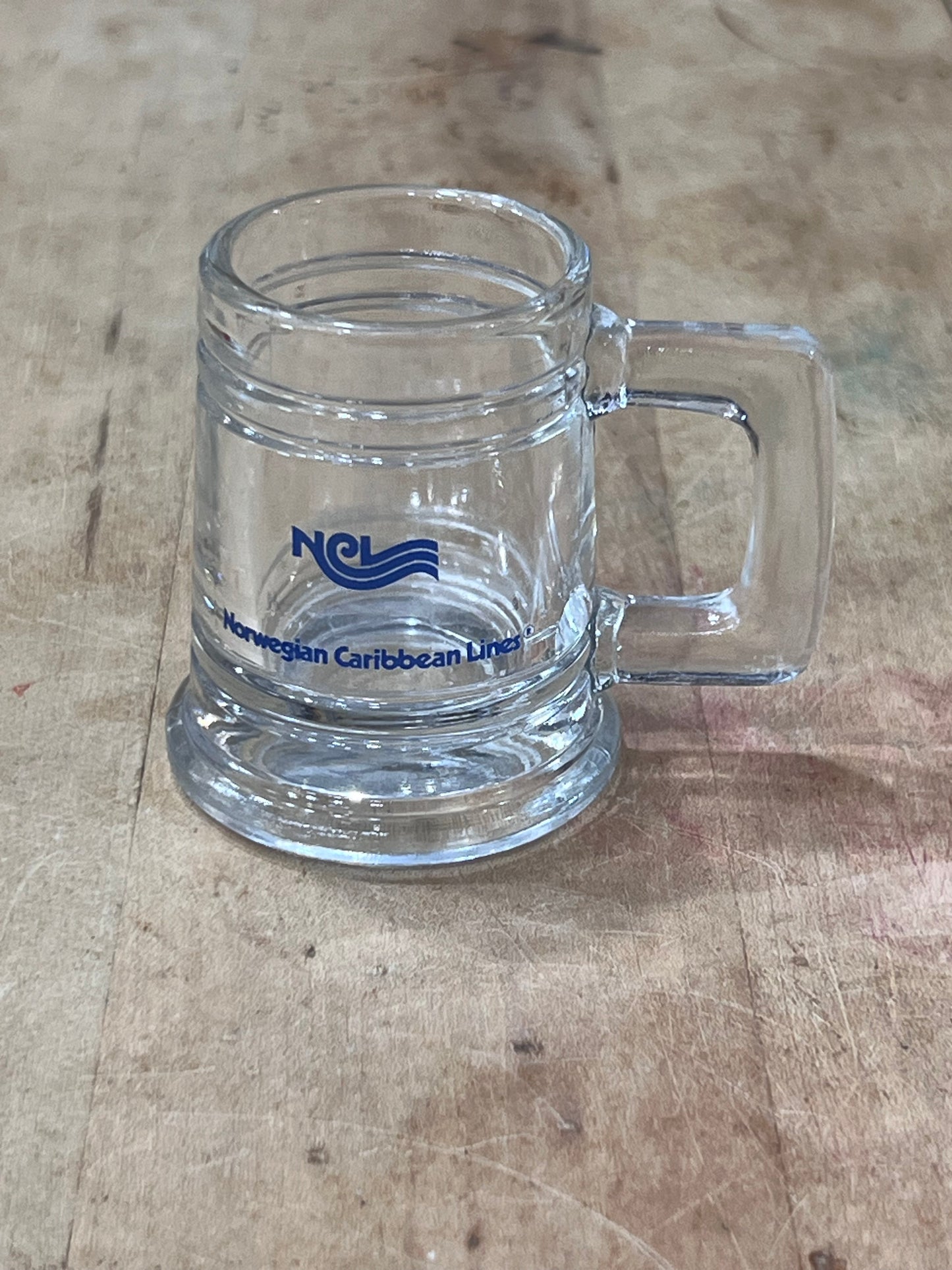 A clear glass beer mug with a handle, featuring a blue printed 'NCL' logo and 'Hawaiian Caribbean Lines' text, resting on a wooden surface.