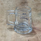 A clear glass beer mug with a thick handle, lying on a worn wooden tabletop. The glass has some blue printed text and logo, but it is not clearly legible.