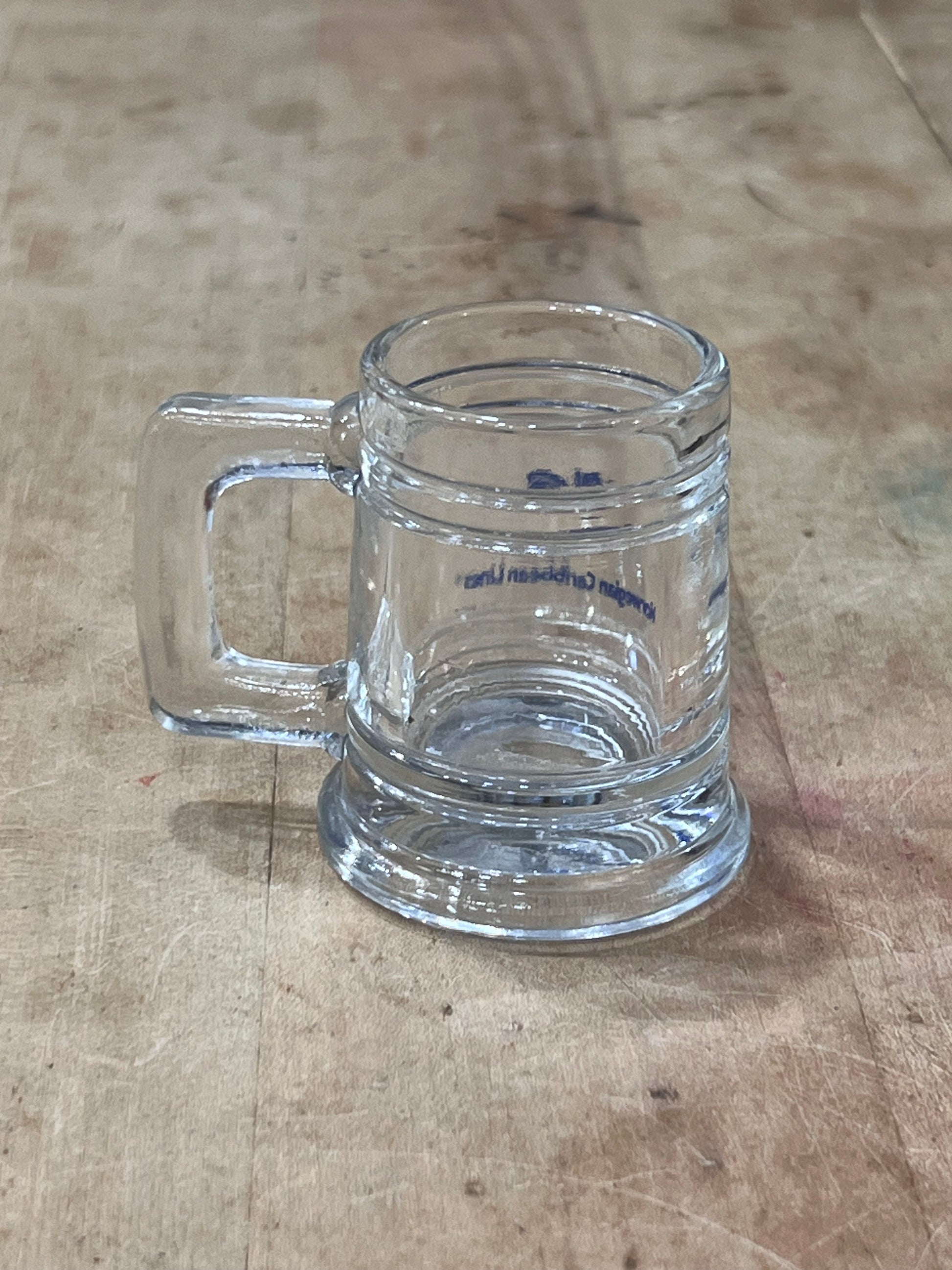 A clear glass beer mug with a thick handle, lying on a worn wooden tabletop. The glass has some blue printed text and logo, but it is not clearly legible.