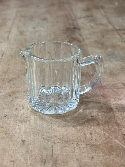A small clear glass creamer pitcher with a handle and spout, featuring a starburst pattern on the base, placed on a rustic wooden surface.