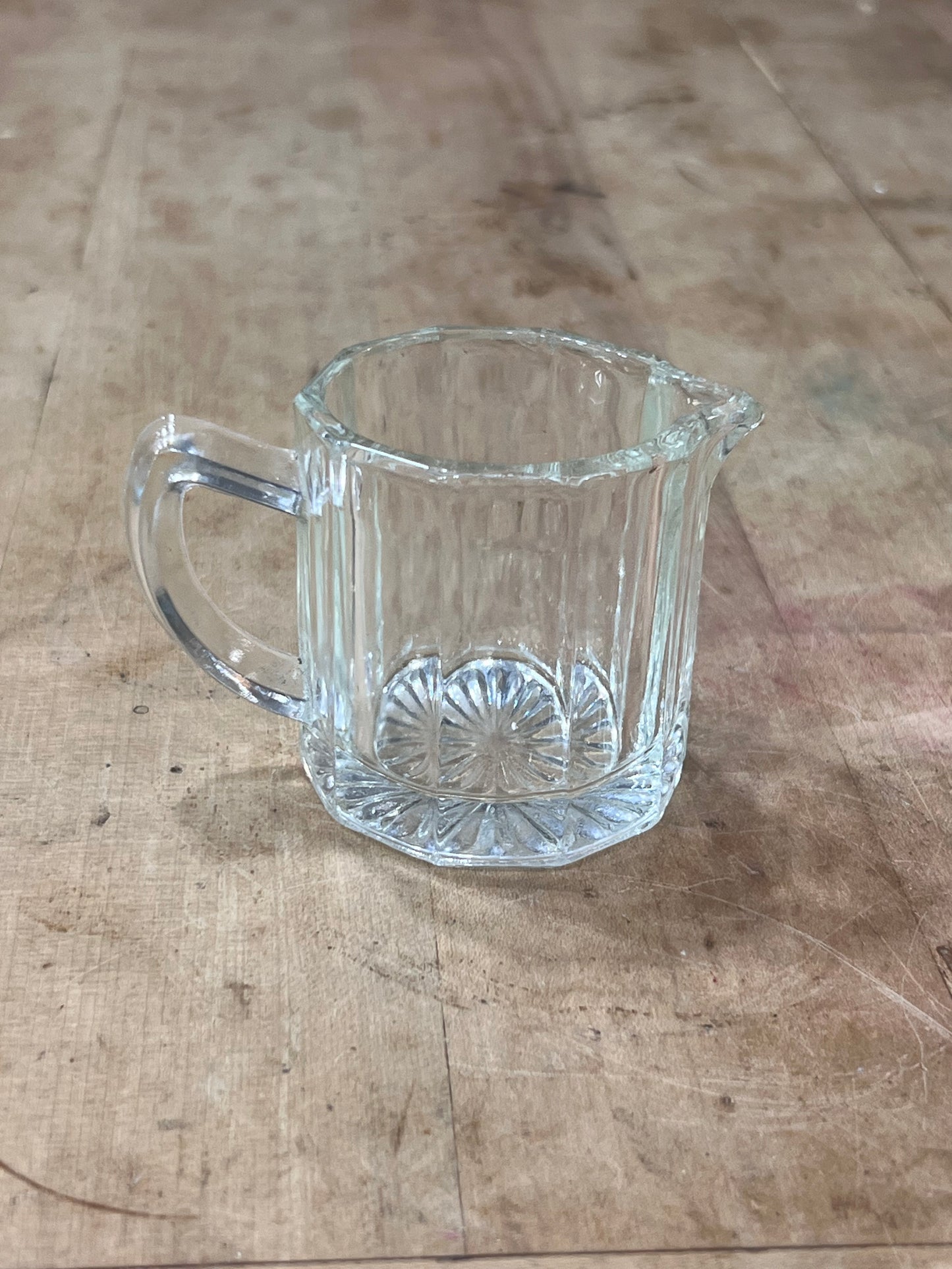 A small clear glass pitcher with a ribbed fluted body, a handle on the left, and a small pouring spout on the right, placed on a rustic wooden tabletop.