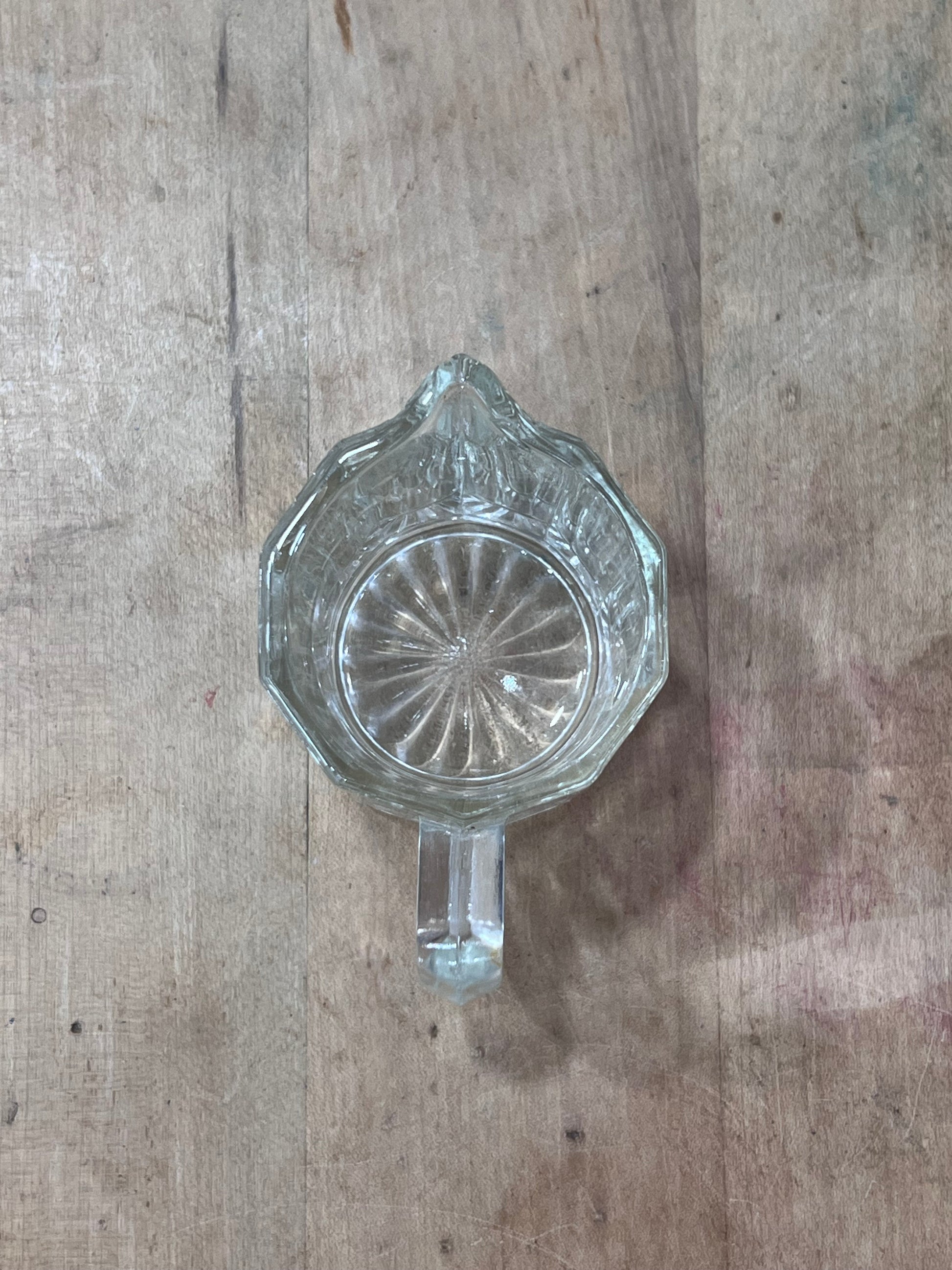 A vintage clear glass pitcher with a faceted, starburst-patterned base and a short handle, shown from a top-down view on a rustic wooden surface.