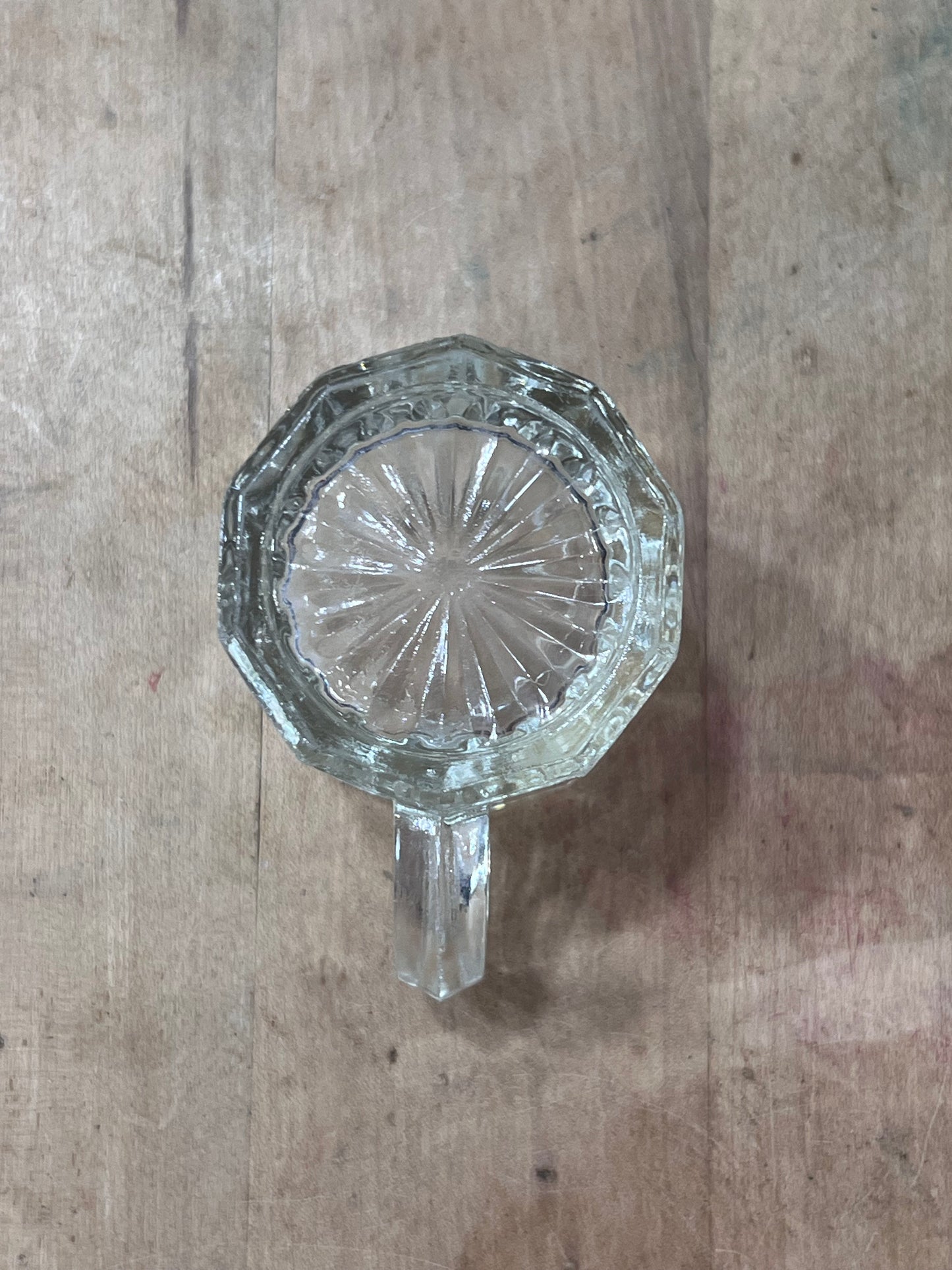 A clear cut-glass mug or tumbler with a faceted octagonal rim and a short handle, photographed from above on a rustic wooden surface.