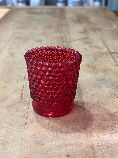 A small red textured glass candle holder or tumbler with a beaded bubble pattern, sitting on a rustic wooden tabletop.