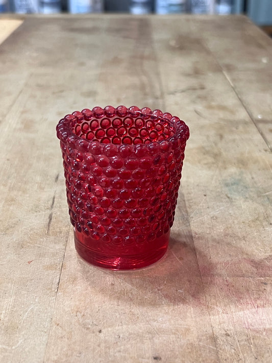 A small red textured glass candle holder or tumbler with a beaded bubble pattern, sitting on a rustic wooden tabletop.