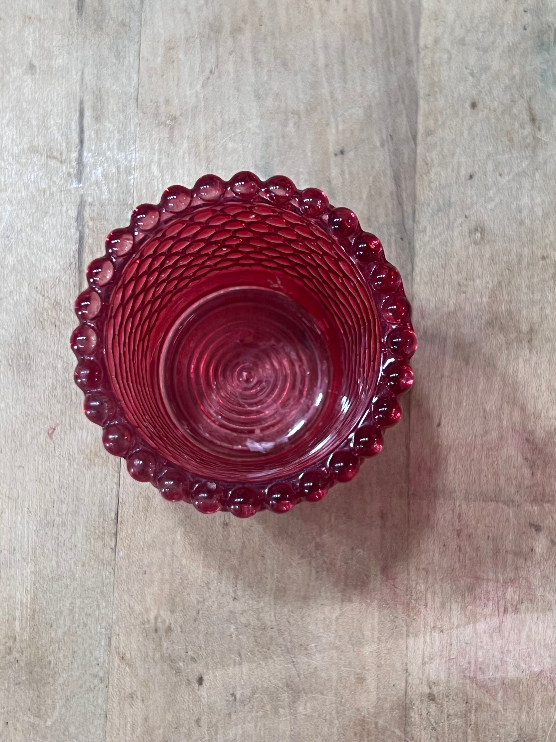 A small round red glass bowl with a textured spiral pattern inside and a scalloped beaded rim, shown from a top-down view on a light wood surface.