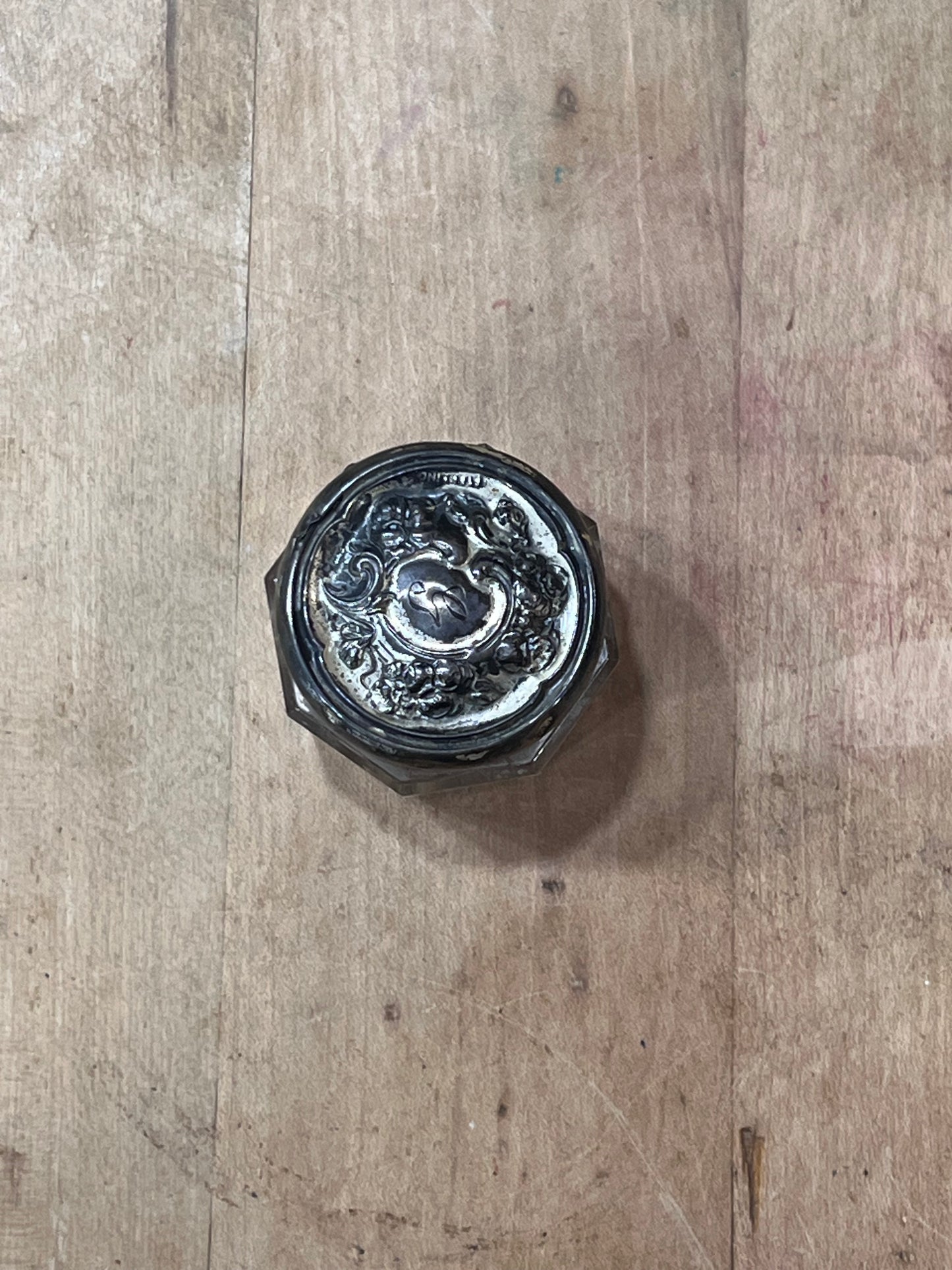 A vintage silver-toned metal jar lid with an embossed floral or scrollwork design, photographed from above on a rustic wooden surface.