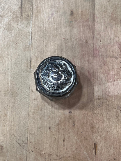 A vintage silver-toned metal jar lid with an embossed floral or scrollwork design, photographed from above on a rustic wooden surface.