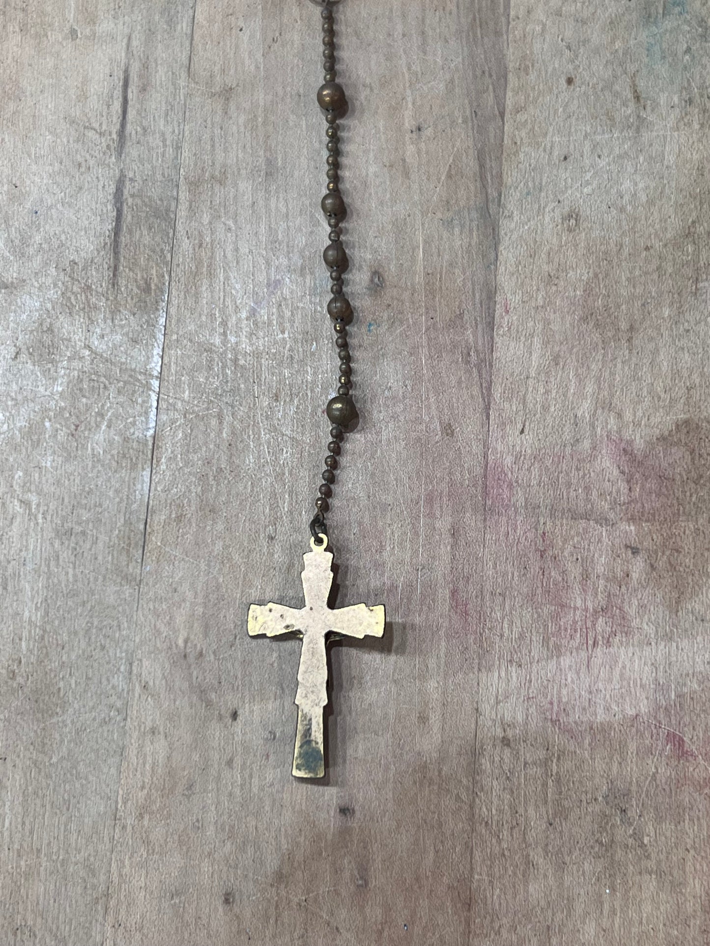 A vintage-style rosary necklace with dark brown metal beads and a distressed, weathered gold cross pendant, laid flat on a rustic wooden surface.