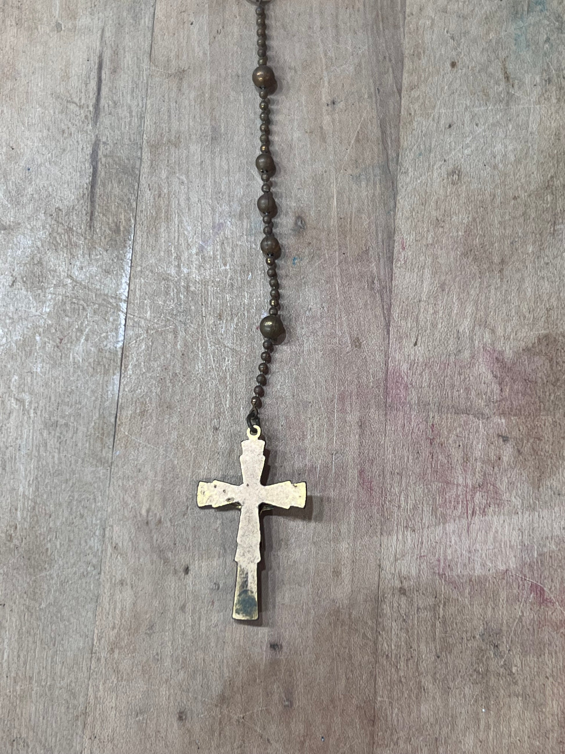 A vintage-style rosary necklace with dark brown metal beads and a distressed, weathered gold cross pendant, laid flat on a rustic wooden surface.
