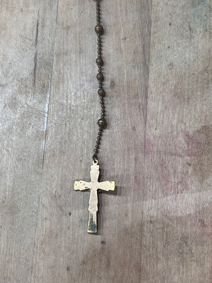 A vintage-style rosary necklace with dark brown metal beads and a distressed, weathered gold cross pendant, laid flat on a rustic wooden surface.