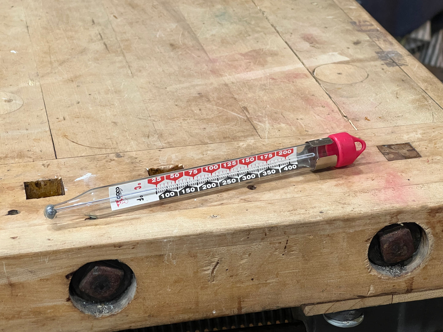 A clear glass thermometer with red and black printed temperature markings and a bright pink cap, resting on a worn wooden workbench.