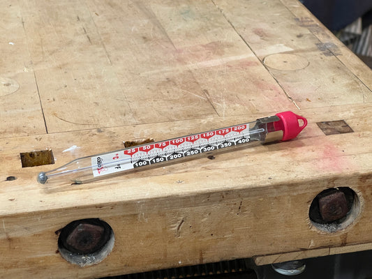 A clear glass thermometer with red and black printed temperature markings and a bright pink cap, resting on a worn wooden workbench.