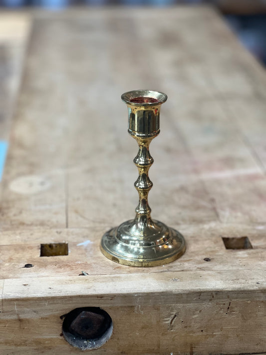 A small brass candlestick holder with a flared base and a cylindrical cup, standing on a wooden workbench.