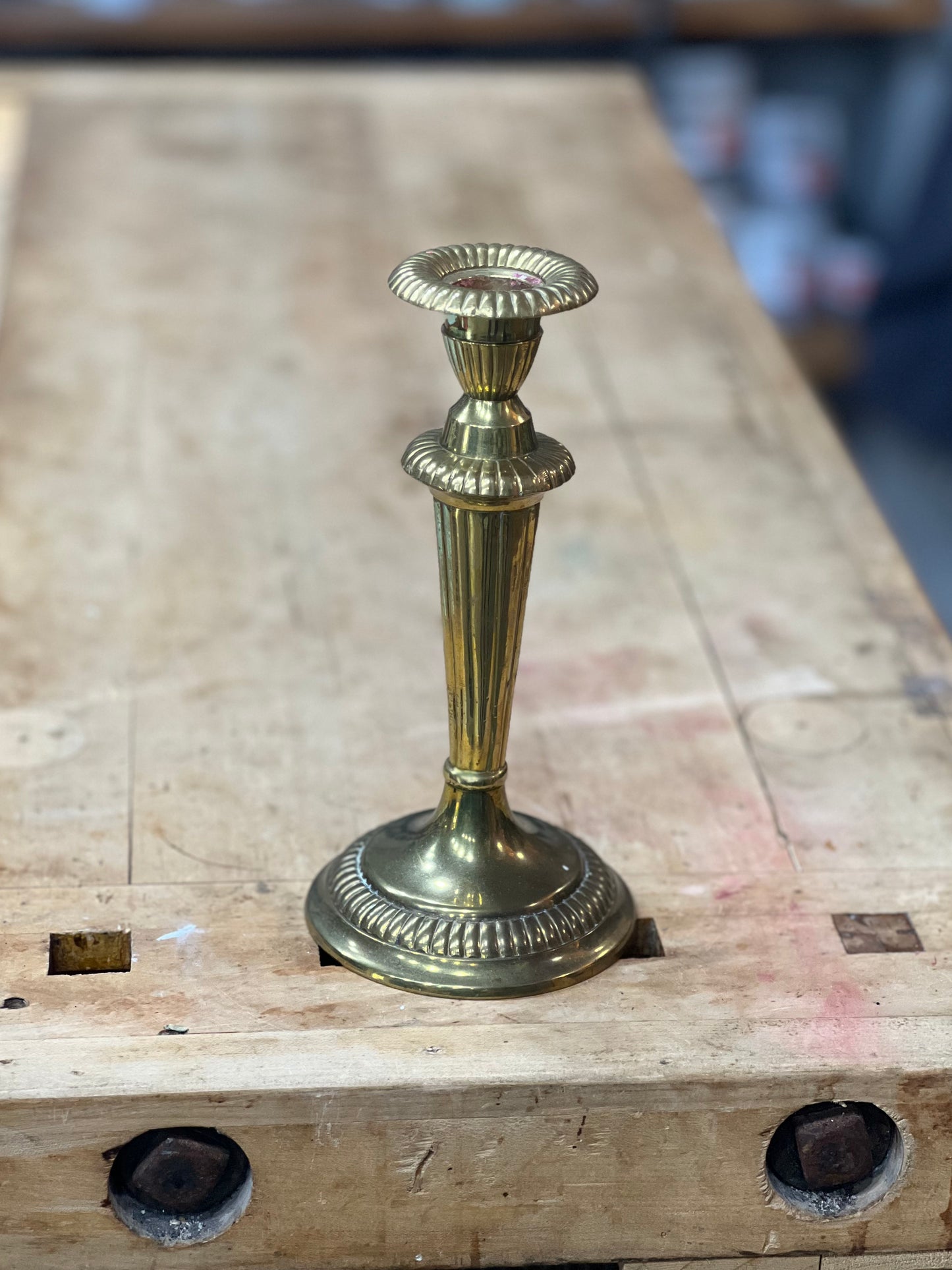 A vintage brass candlestick holder with a fluted stem and circular base, standing on a wooden workbench.