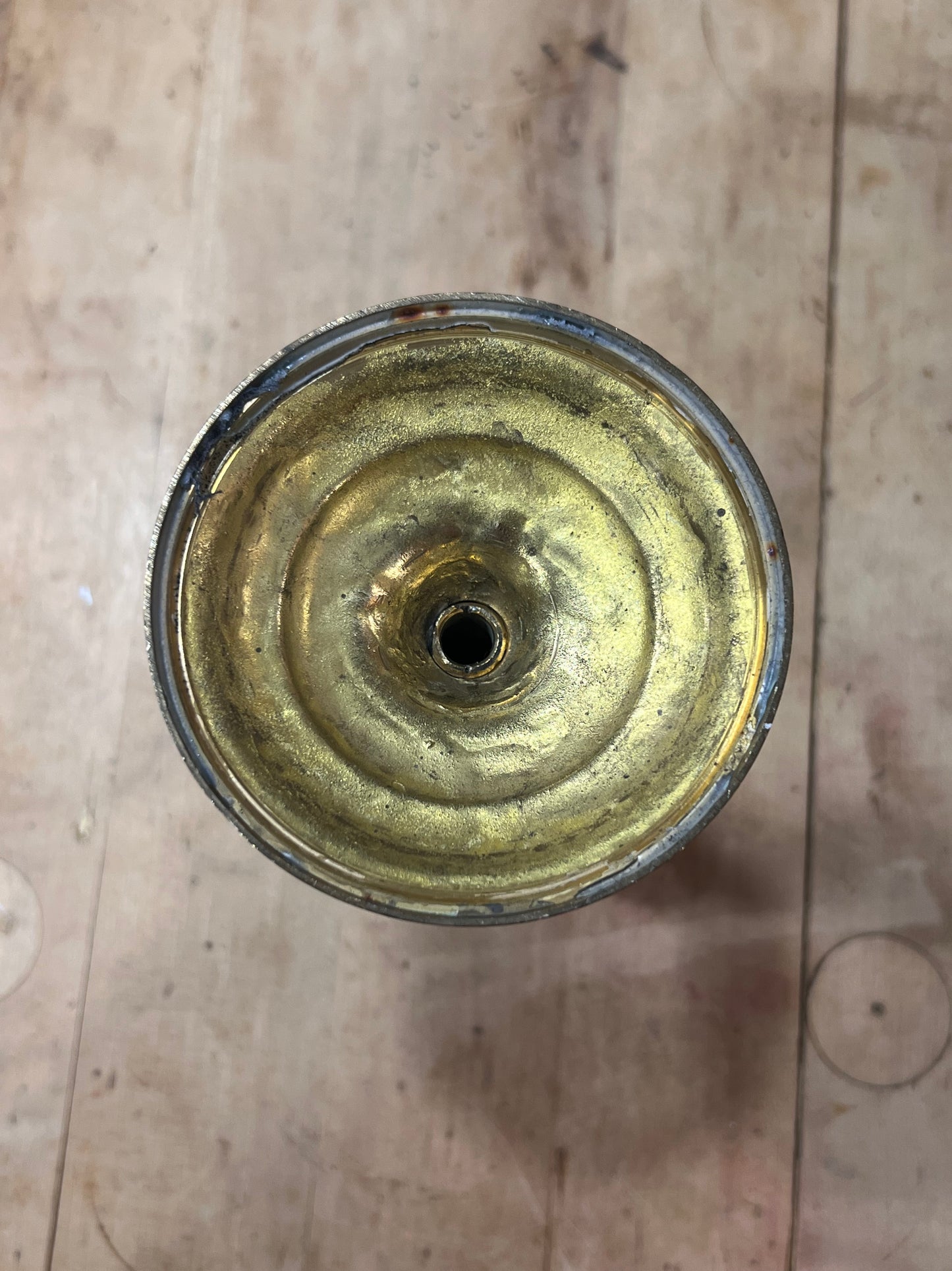 A top-down view of a round, metallic gold-colored object with concentric circular ridges and a small hole in the center, resting on a light-colored wooden surface.