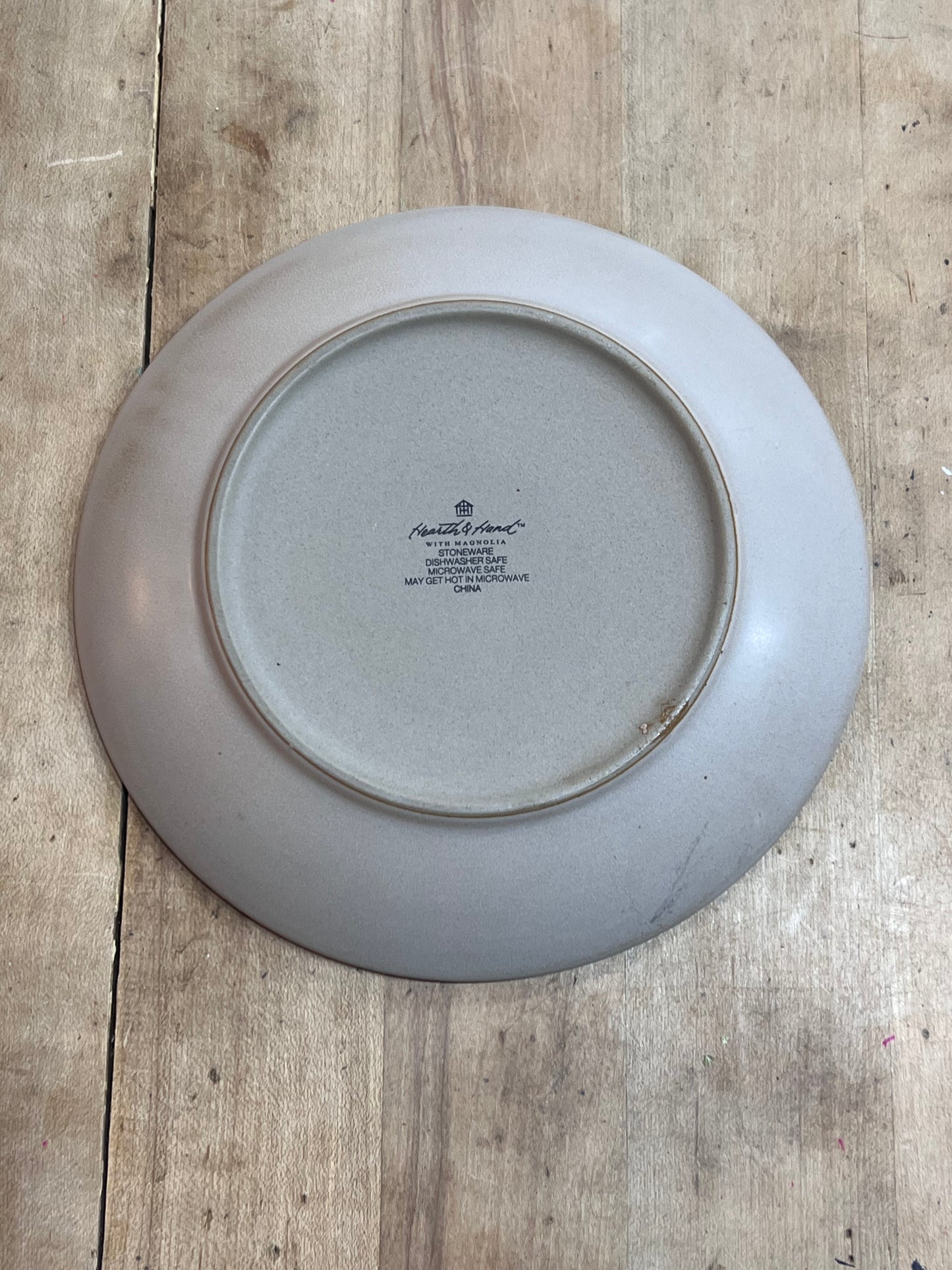 The underside of a round ceramic plate with a light gray glaze, featuring the printed text 'Royal Albert' and 'The Queen Mother Honored Edition 1974', placed on a rustic wooden surface.
