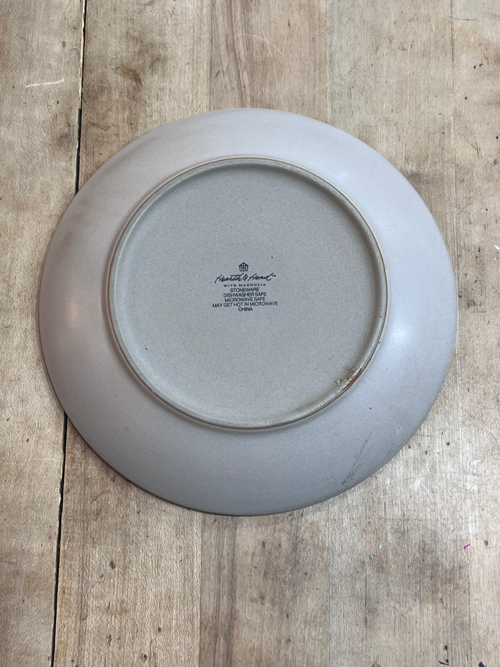 The underside of a round ceramic plate with a light gray glaze, featuring the printed text 'Royal Albert' and 'The Queen Mother Honored Edition 1974', placed on a rustic wooden surface.